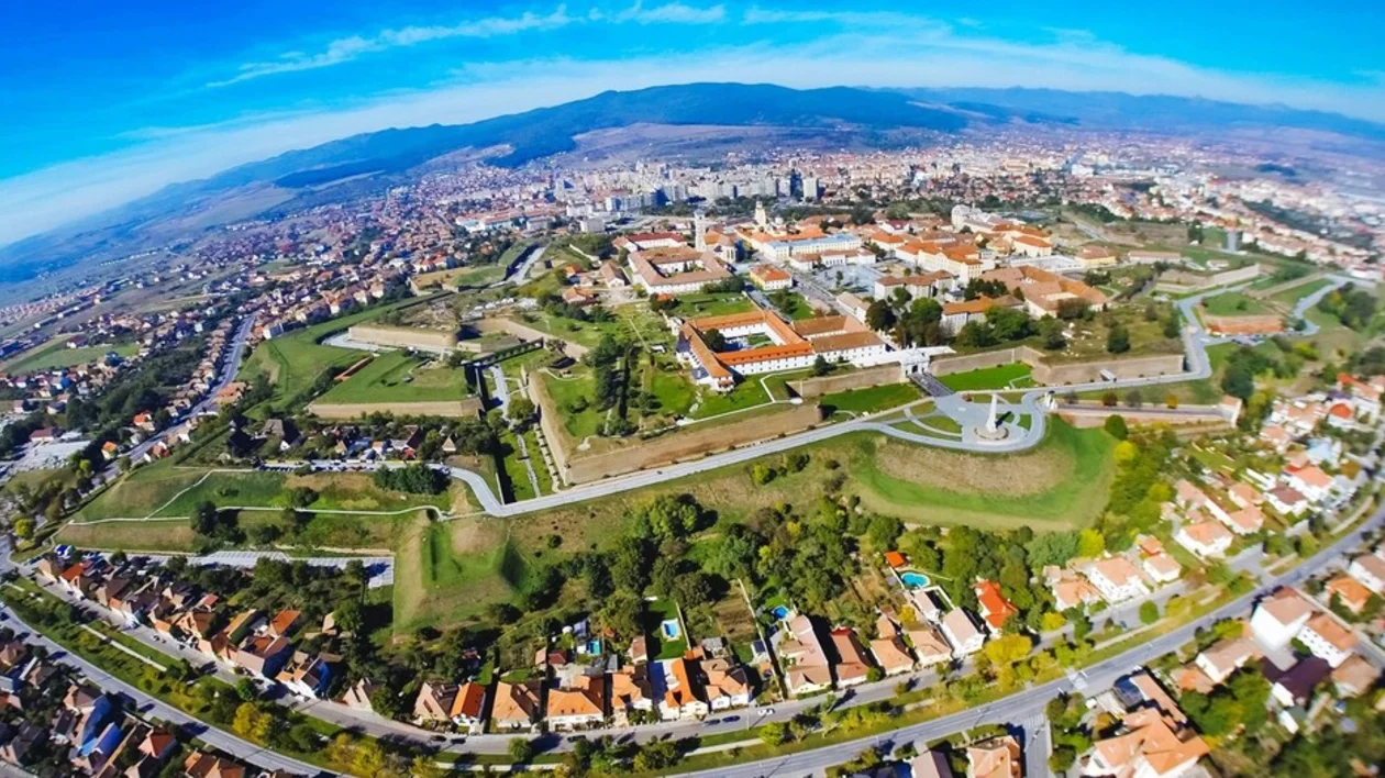 Aerial view of a city featuring green spaces, historic buildings, and residential neighborhoods against a backdrop of mountains and blue sky.