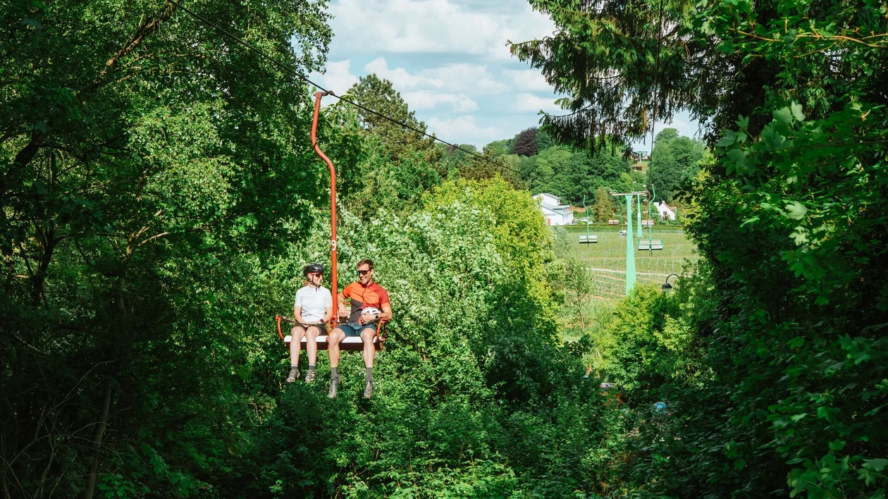 Two people on a chairlift above green vineyards and forest.