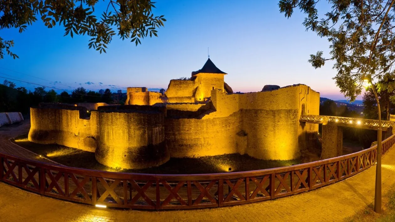 Illuminated medieval castle at twilight, surrounded by trees, with a wooden walkway and a clear sky transitioning to night.