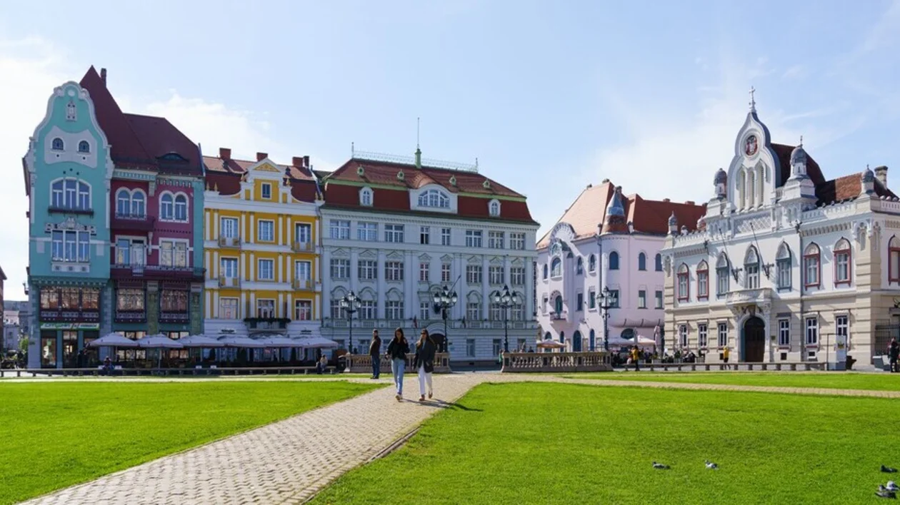 Colorful historic buildings line a grassy square, with people walking on a path under a bright blue sky.
