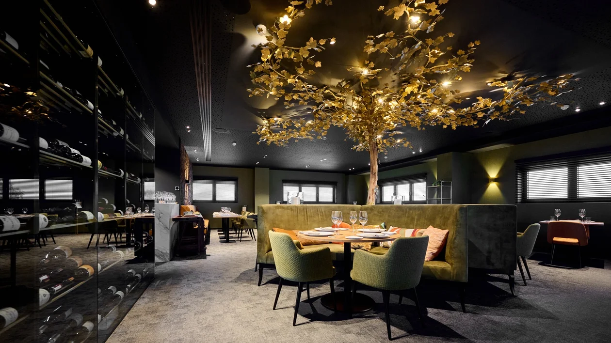 Elegant dining room with soft green seating, wine racks along the wall, and a golden tree installation with illuminated leaves as a central ceiling decoration.