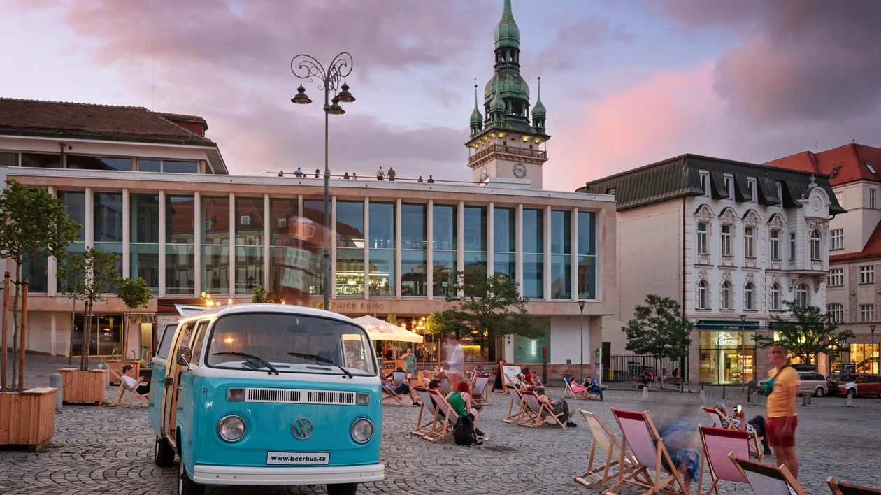 Retro blue VW van and people relaxing on deck chairs at Zelný Market in Brno during a summer evening, with the old town hall tower in the background.