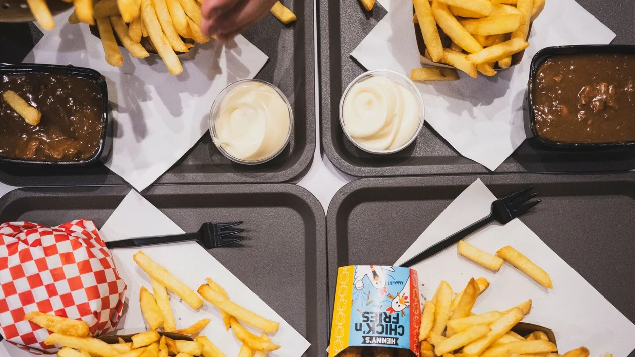 A top-down view of four trays filled with French fries, a burger, sauces, and chicken tenders, with a hand pouring more fries.