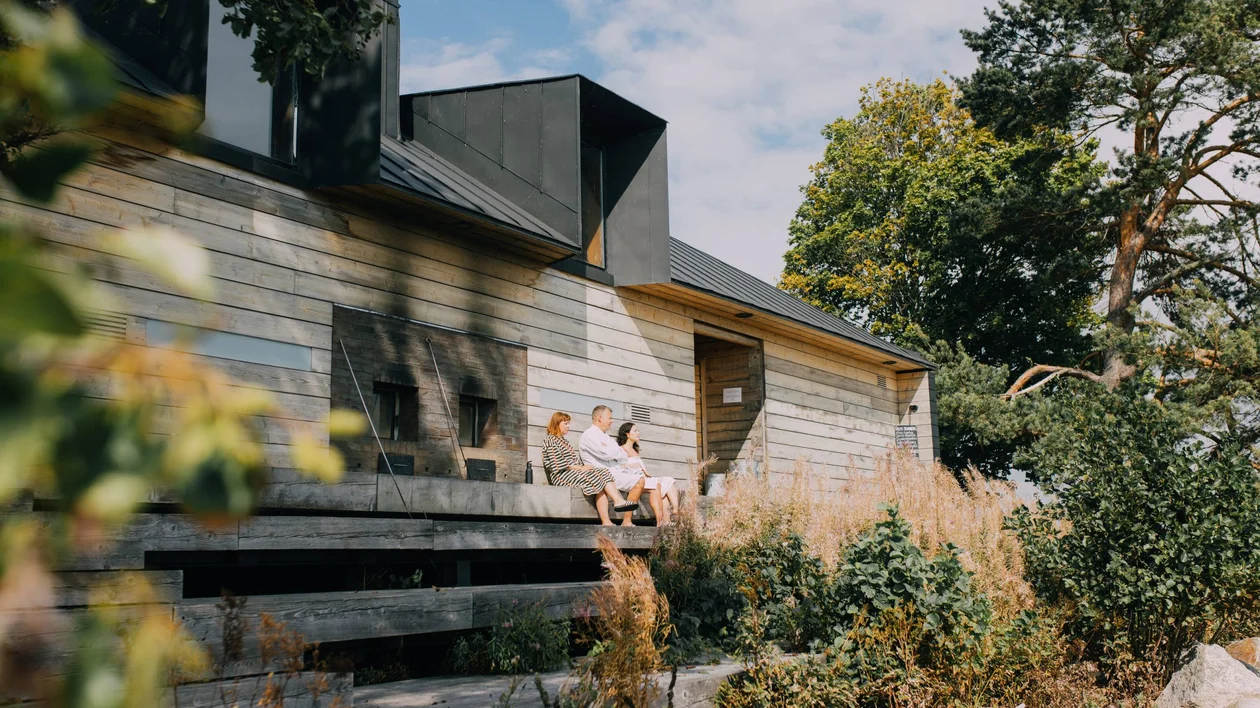A modern wooden house with a sloped black roof, surrounded by greenery, where a group of people enjoys a sunny day on the steps.