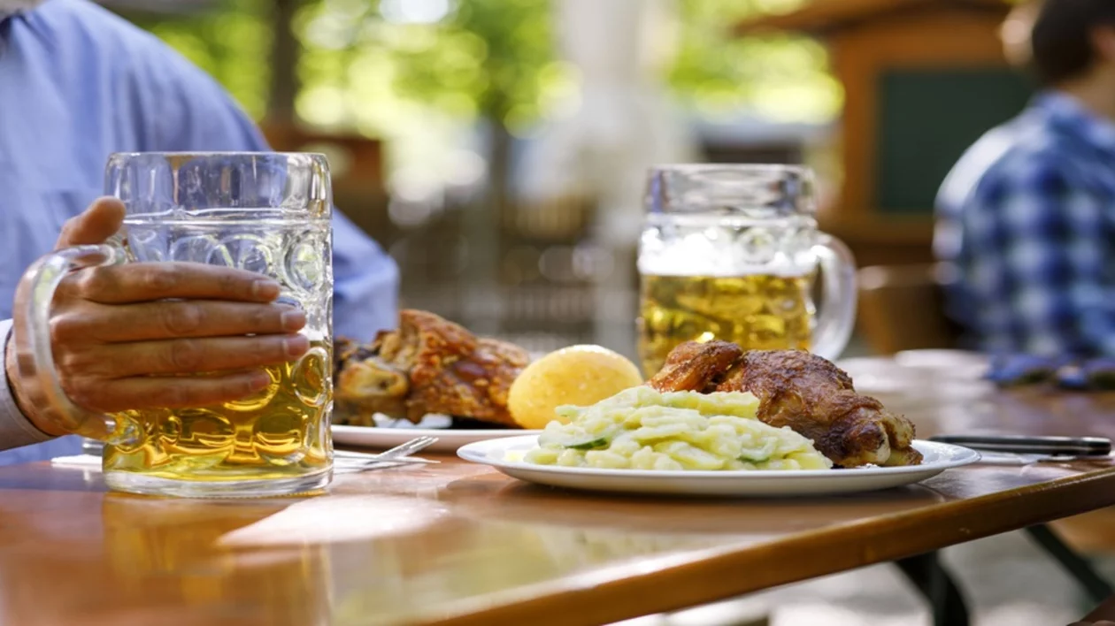 A hand holds a large beer mug beside a plate of roasted chicken, creamy mashed potatoes, and a lemon slice on a sunny outdoor table.