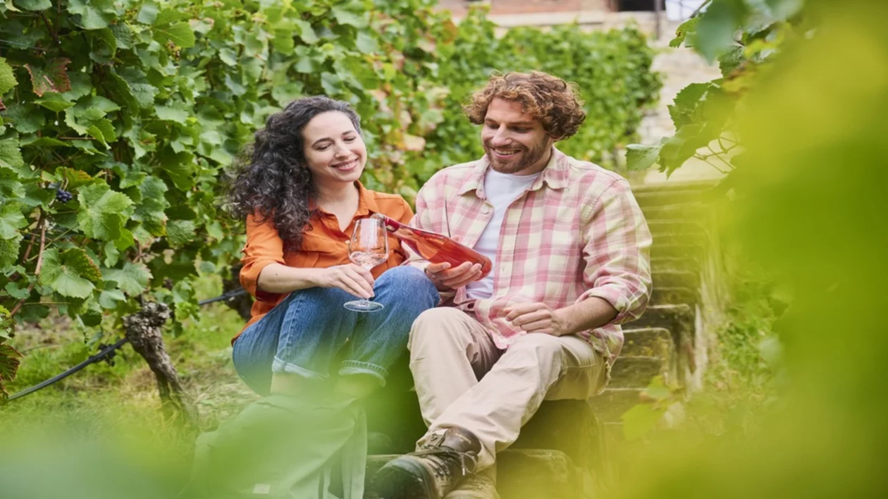 A couple enjoys wine together in a vineyard, seated on stone steps surrounded by lush green grapevines.
