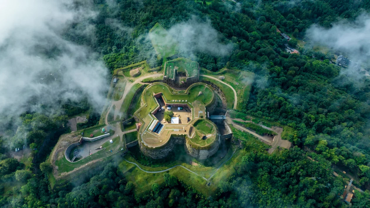 Srebrna Gora Castle seen from above, surrounded by dense green scenery and a cloudy sky backdrop.