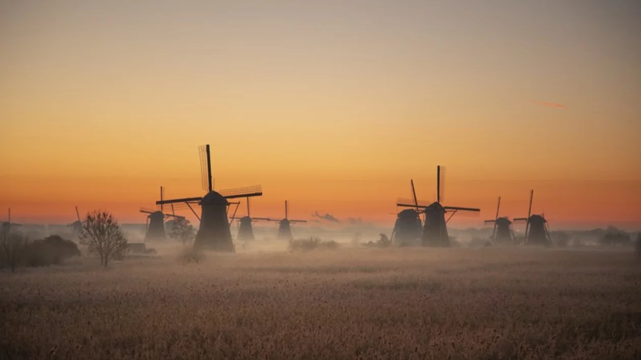 Sunset view of the windmills with clouds between them, painted in orange, yellow, and blue hues, with dry brown grass in the foreground.