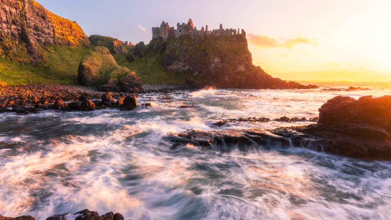 Dunluce Castle perched dramatically on a rugged cliff in County Antrim, Northern Ireland, with the Atlantic Ocean crashing below and a vibrant sunset sky above.