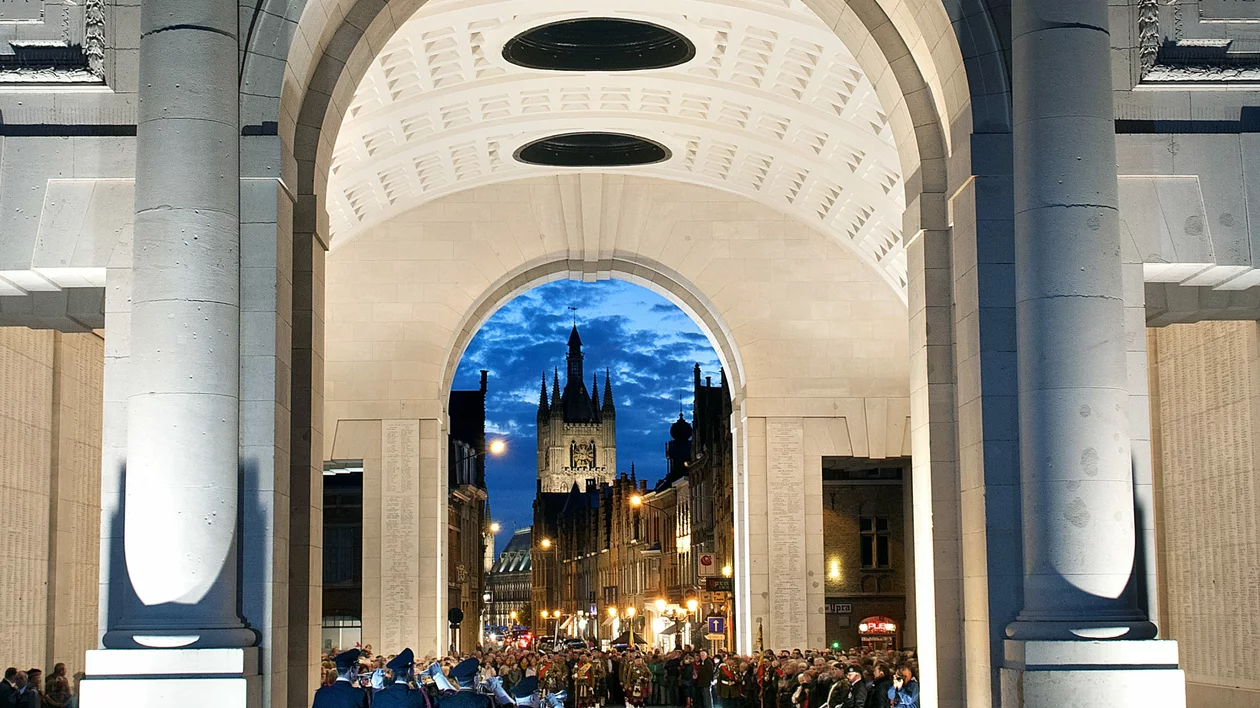 Night scene of soldiers playing trumpets during The Last Post ceremony, illuminated by lights, with a cathedral in the background and people watching.