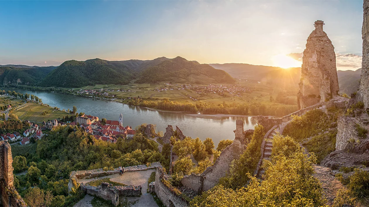 A high-angle panoramic sunset view of the Danube River valley, showing the red-roofed village of Dürnstein nestled by the water, framed by the sun-drenched stone ruins of Dürnstein Castle in the foreground.