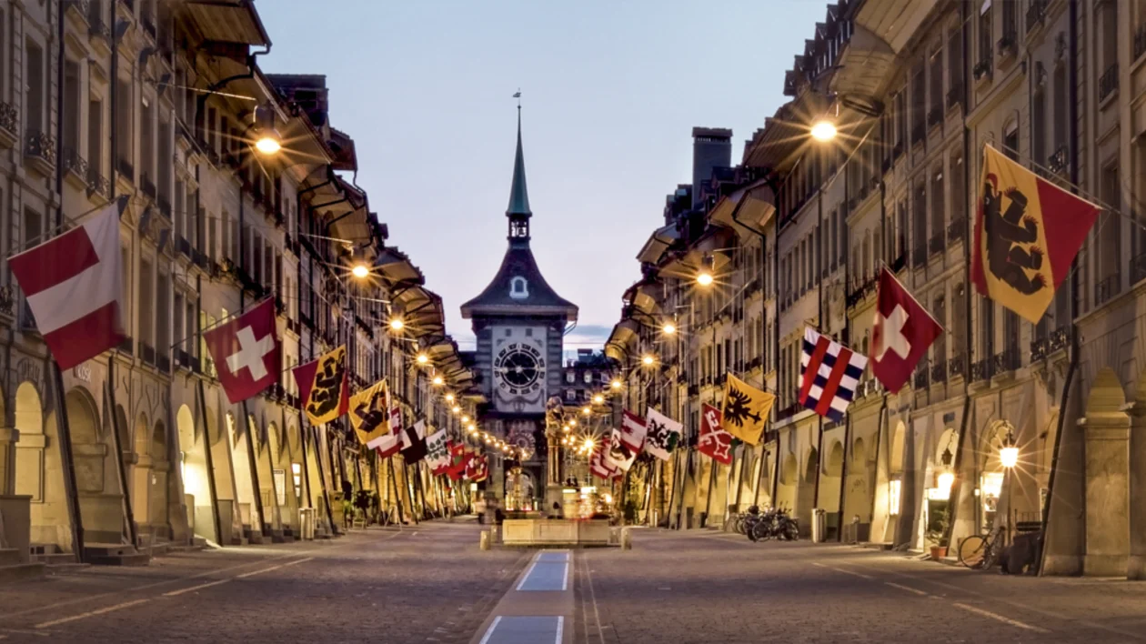 Historic street in Bern, Switzerland, decorated with Swiss and cantonal flags, leading to the iconic Zytglogge clock tower at dusk.