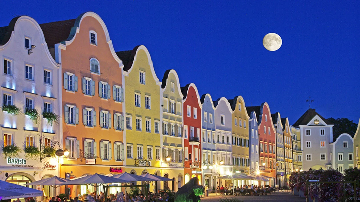 Lively plaza in Schärding at night, featuring colorful Baroque architecture and a glowing full moon.