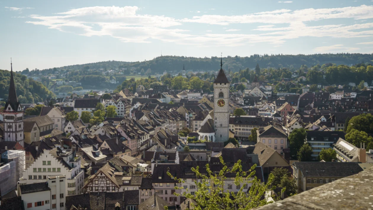 View of the old town of Schaffhausen on a sunny day, with white buildings, a clock tower, a church, and green hills in the background.