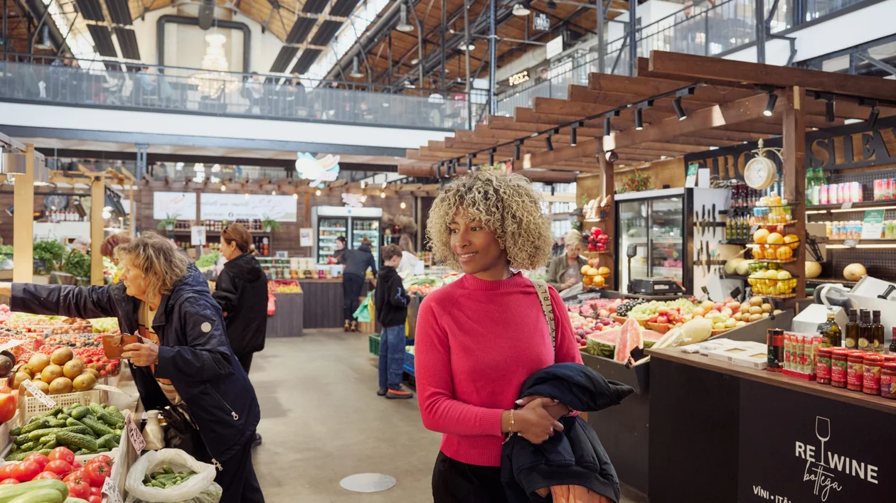 A woman browsing fresh produce stalls inside Riga’s Āgenskalns Market, a lively indoor food market with wooden structures and high ceilings.