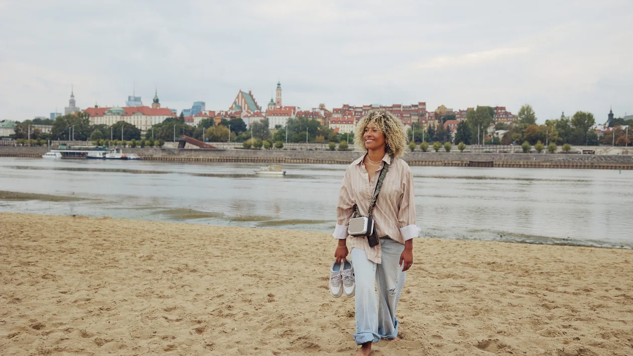 Woman walking barefoot on a sandy beach by the Vistula River with Warsaw’s Old Town skyline in the distance.