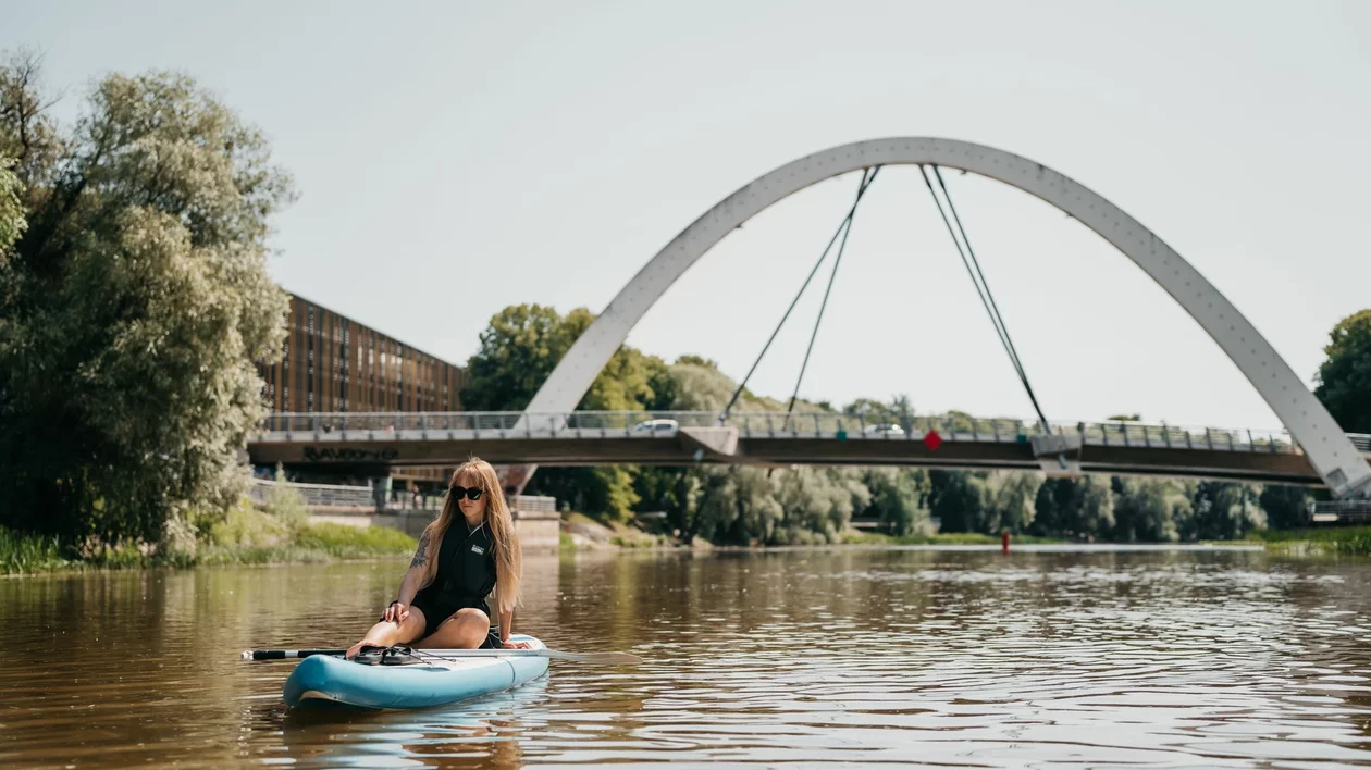 Woman paddleboarding (SUP) on the Emajõgi River in Tartu, Estonia, with the Kaarsild arch bridge in the background.
