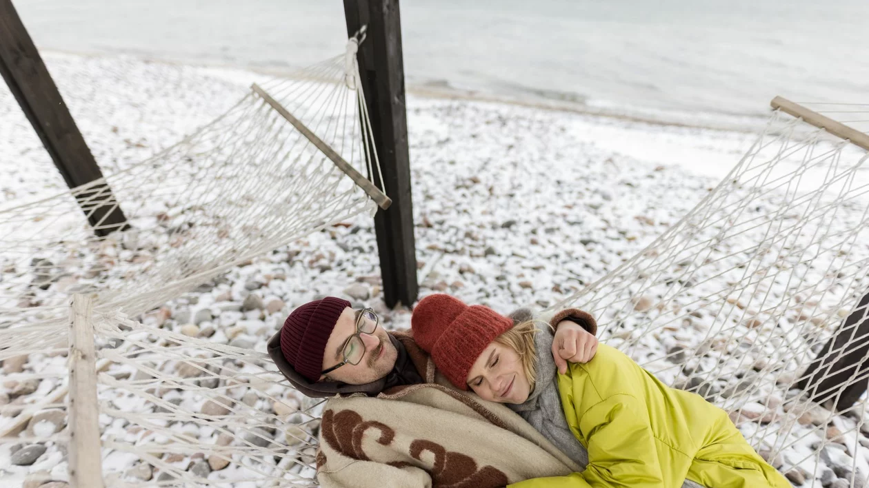 A couple wrapped in a blanket relaxing together in a hammock on a snowy, rocky beach.