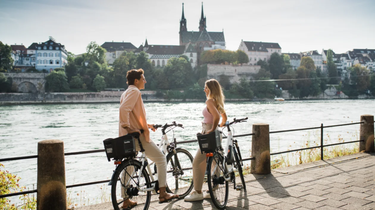 Two cyclists facing each other on a sunny day in Basel, Switzerland, with the Rhine River and historic buildings in the background.