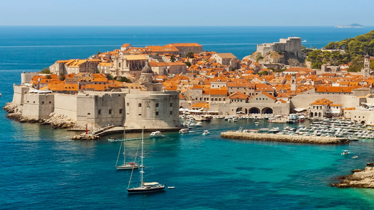 Aerial view of the medieval city walls, historic port, and terracotta roofs of Dubrovnik, Croatia, overlooking the turquoise Adriatic Sea.