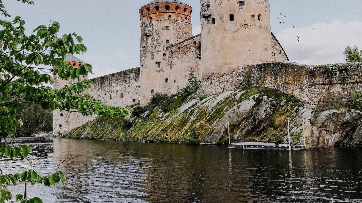 Girl sitting on a waterfront platform facing Olavinlinna Castle in Savonlinna, Finland, surrounded by calm lake waters and forested shoreline.