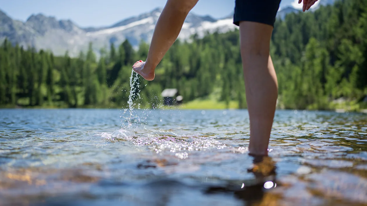 Close-up of a person wading and splashing in a clear Alpine lake with snow-capped mountains in the background.