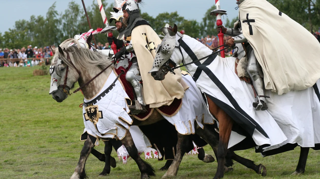 Knights in full armour and white cloaks riding armoured horses during a historical battle reenactment, likely the Battle of Grunwald.