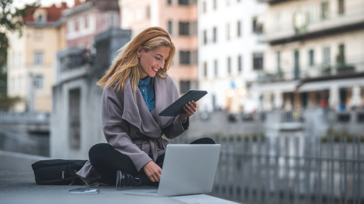 A woman sitting outdoors in a European city, working on a laptop and holding a tablet while smiling.
