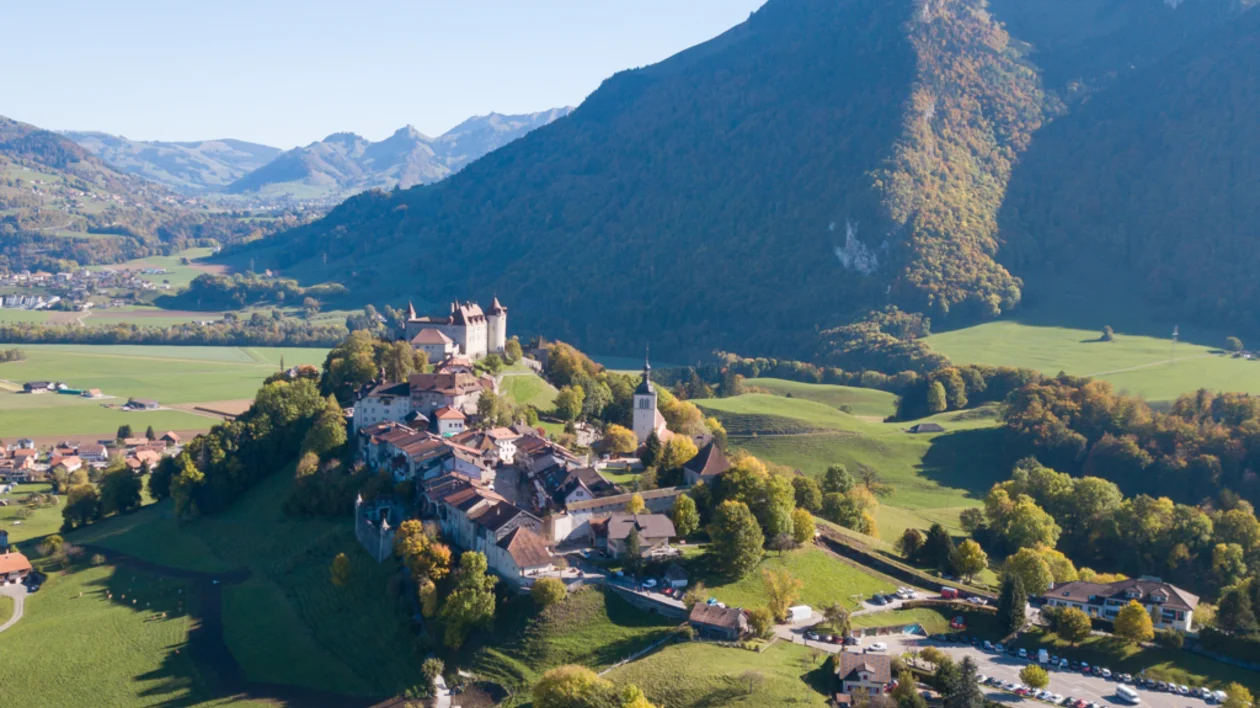 Hilltop village with a castle, surrounded by green fields and mountains under a clear sky.