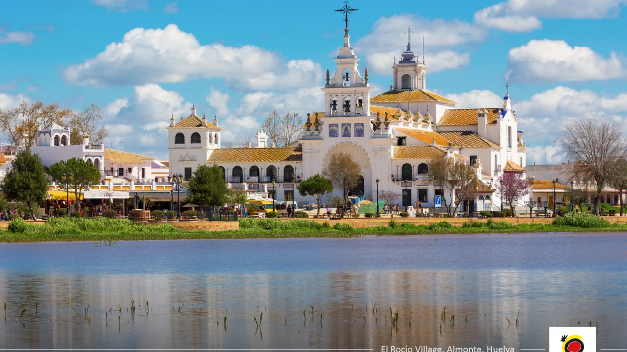 White church with towers and cross reflected in a calm lake under a blue sky with clouds.