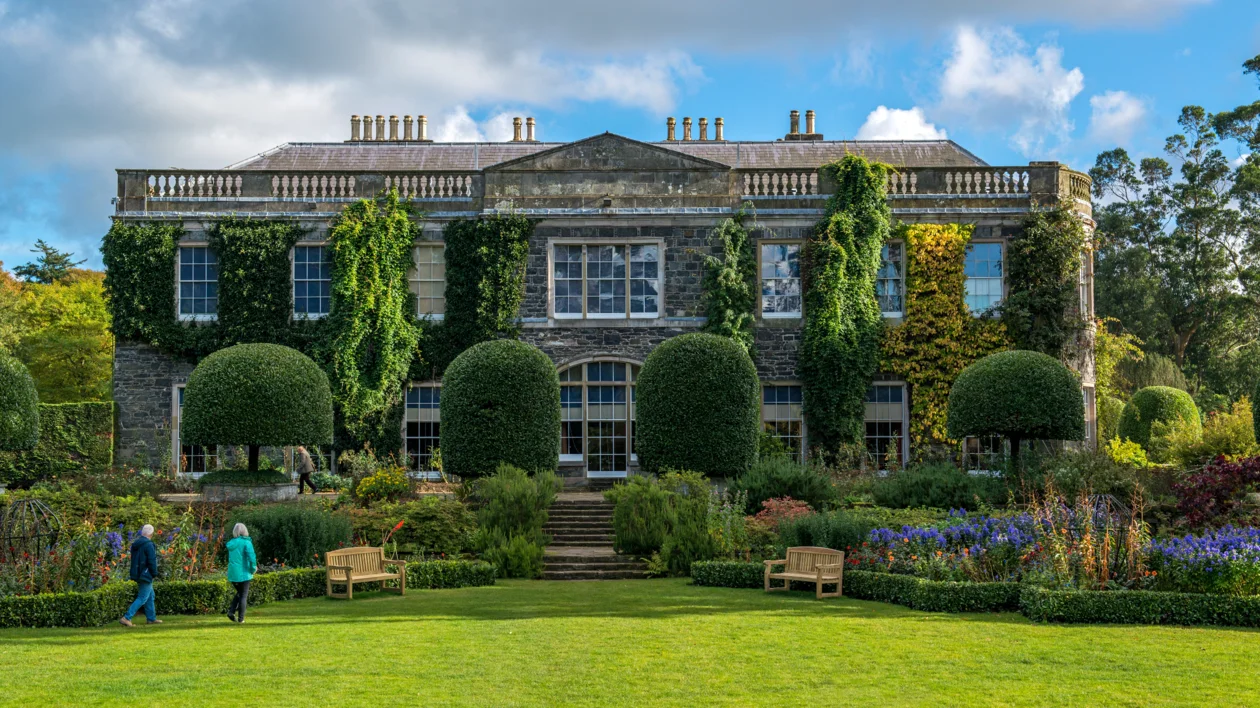 Historic mansion with ivy-covered walls and manicured gardens under a partly cloudy sky.