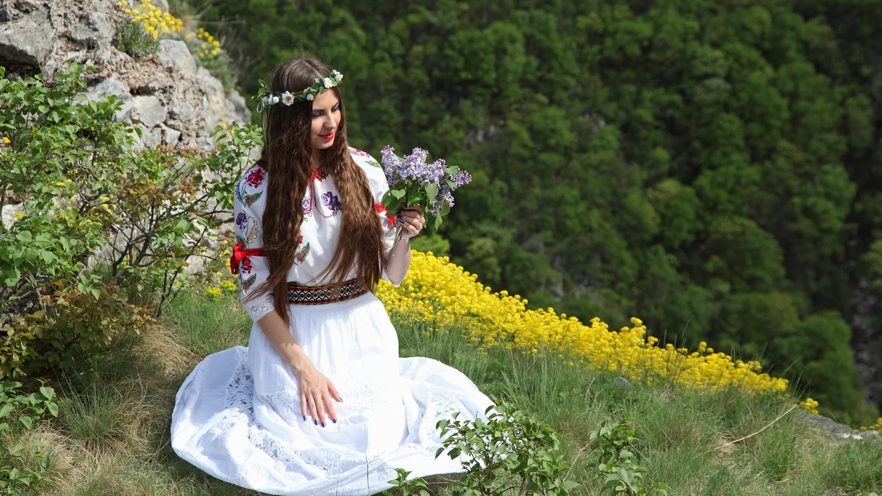 Woman in traditional dress kneeling on grassy hillside, holding flowers, surrounded by nature.