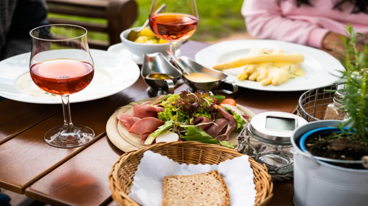 Outdoor table with wine, salad, bread, and plates of food, two people partially visible.