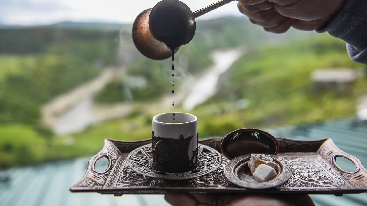 Coffee being poured into a small cup on a tray with sugar cubes, green hills in the background.