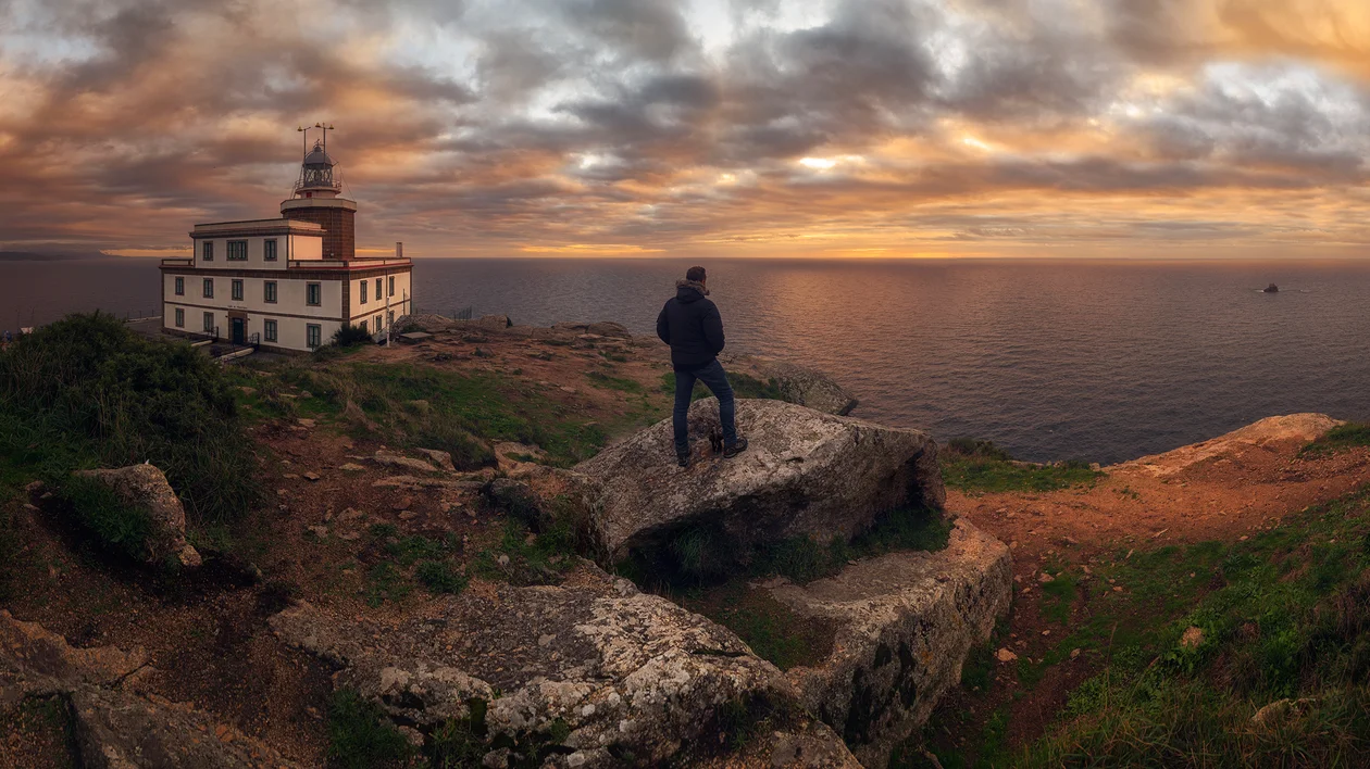 Person standing on rocks near a lighthouse at sunset overlooking the sea.