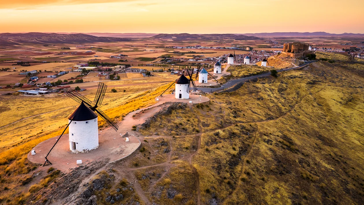 Row of historic windmills on a hill at sunset with fields and a village in the background.