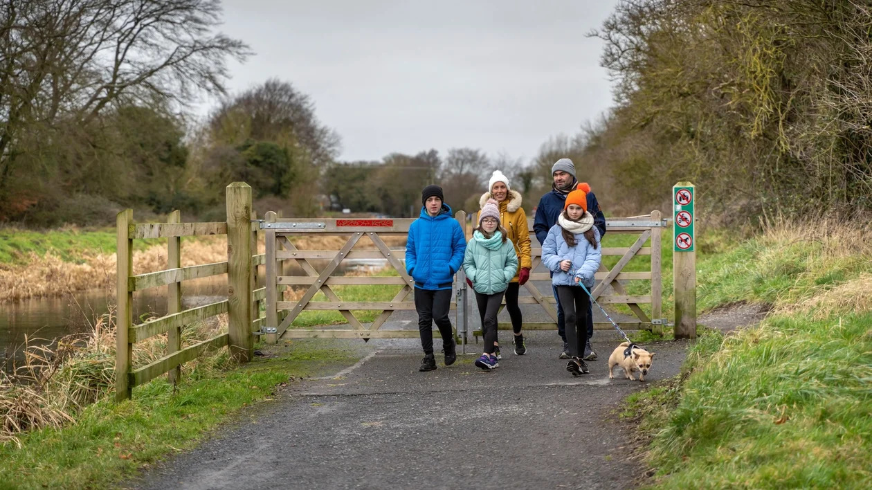 Family and small dog walking on a country path near a wooden gate on a cloudy day.