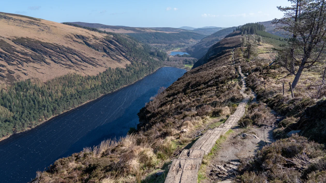 A winding path runs along a lakeside on a sunny hilltop, surrounded by trees and valleys.