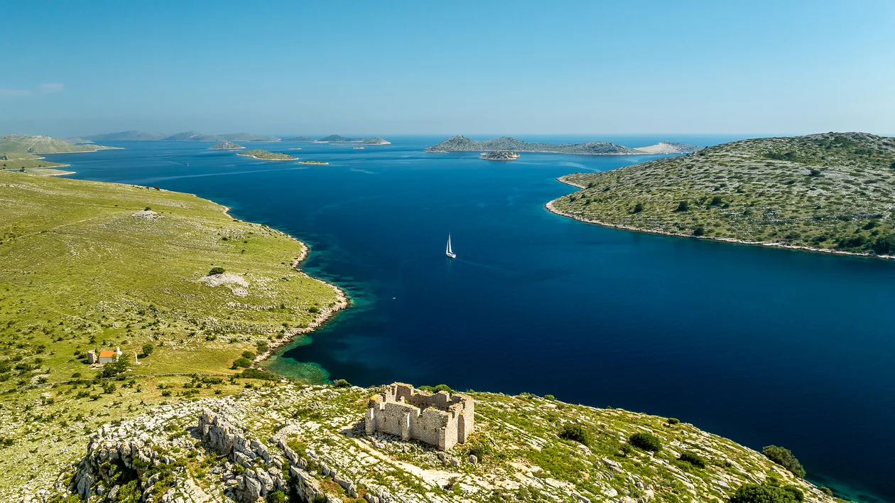 Sailboat on deep blue bay surrounded by green hills and scattered islands under clear sky.