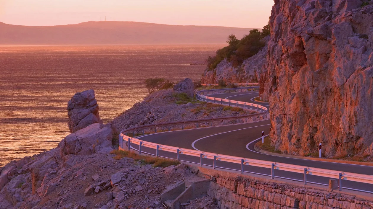 Winding coastal road with guardrails beside a rocky cliff at sunset.