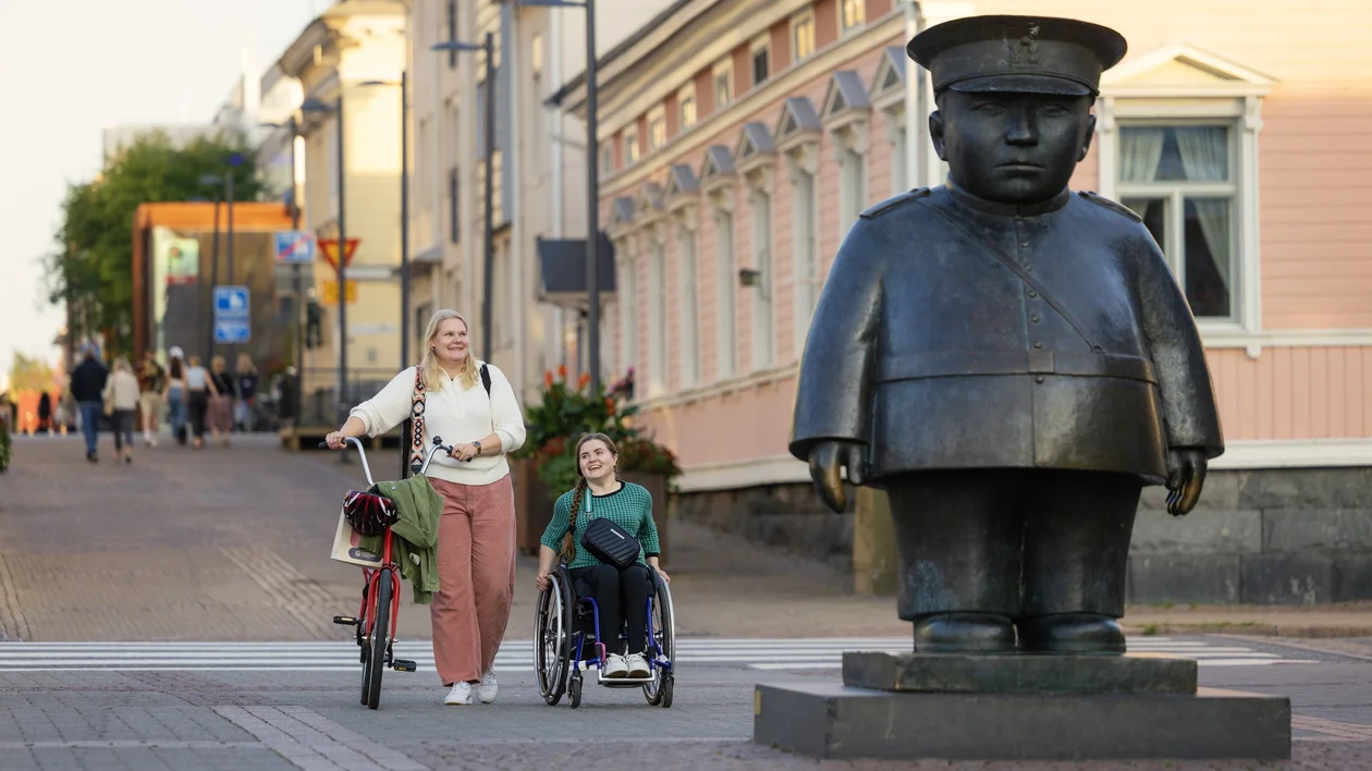 Woman with bicycle and person in wheelchair beside a large statue on a city street.