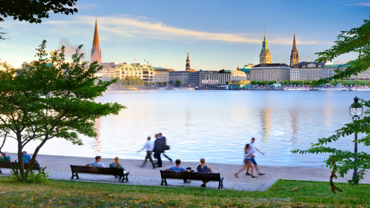 People walking and sitting by a riverside park with city skyline and spires in the background.