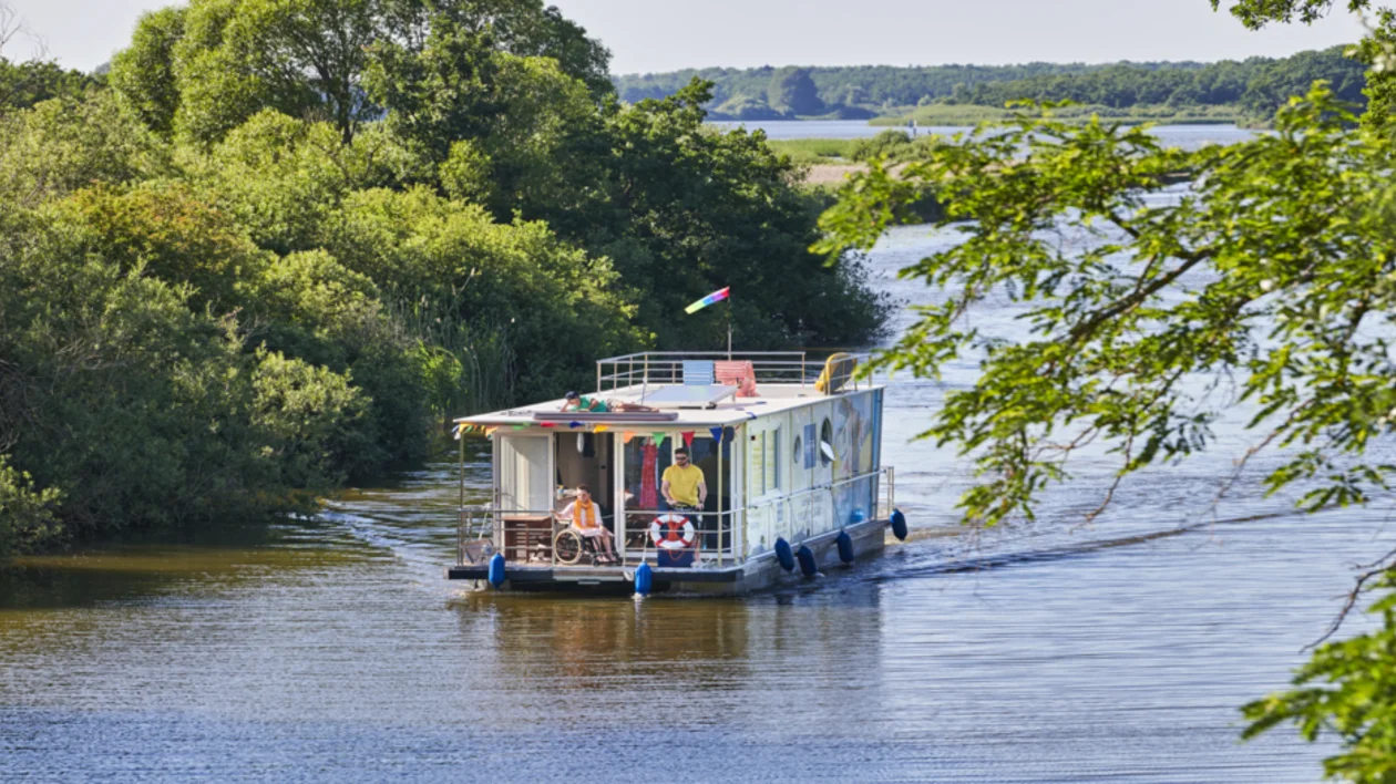 Houseboat with colorful flags moving along a calm river surrounded by green trees.