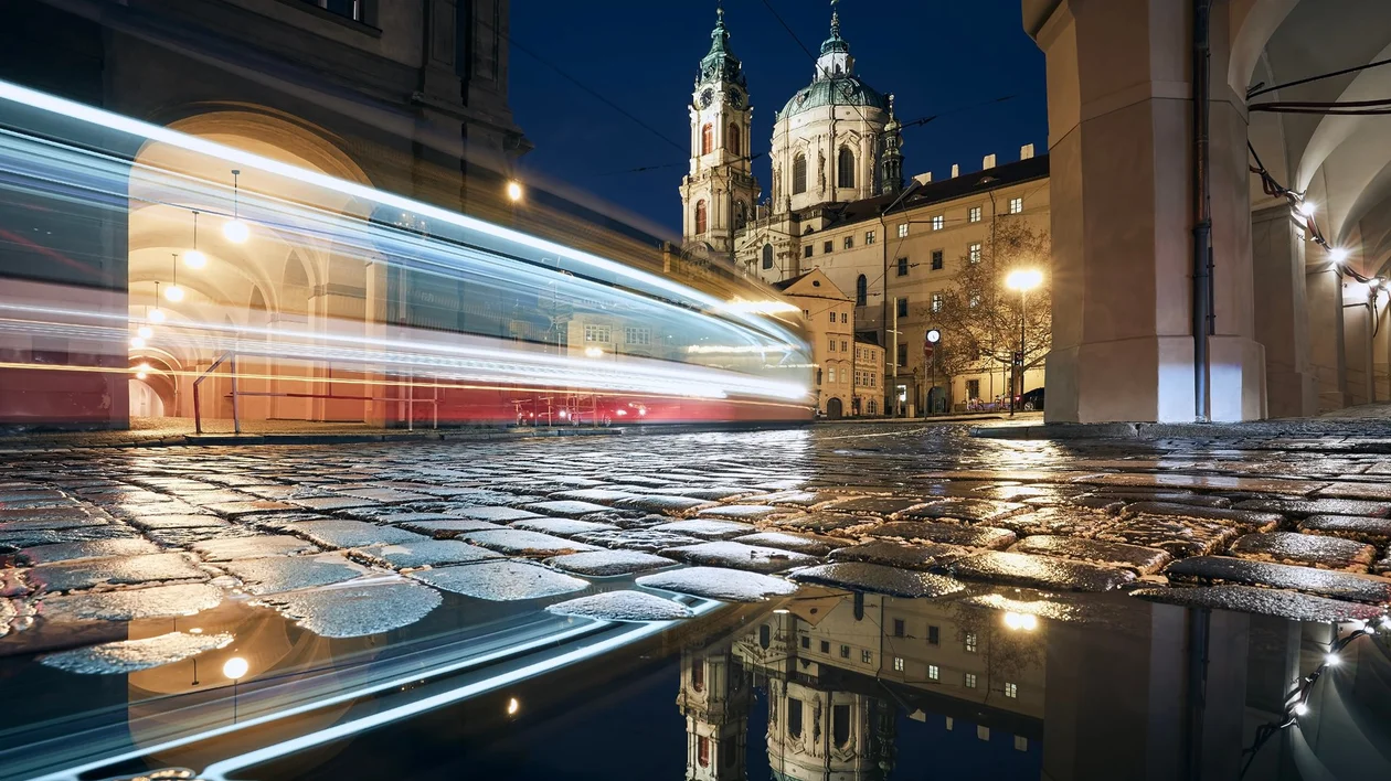 Night city street with blurred lights, wet cobblestones, and illuminated church towers.