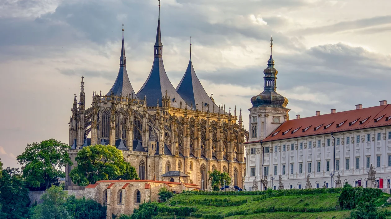 Gothic cathedral with tall spires beside historic white buildings under a cloudy sky.