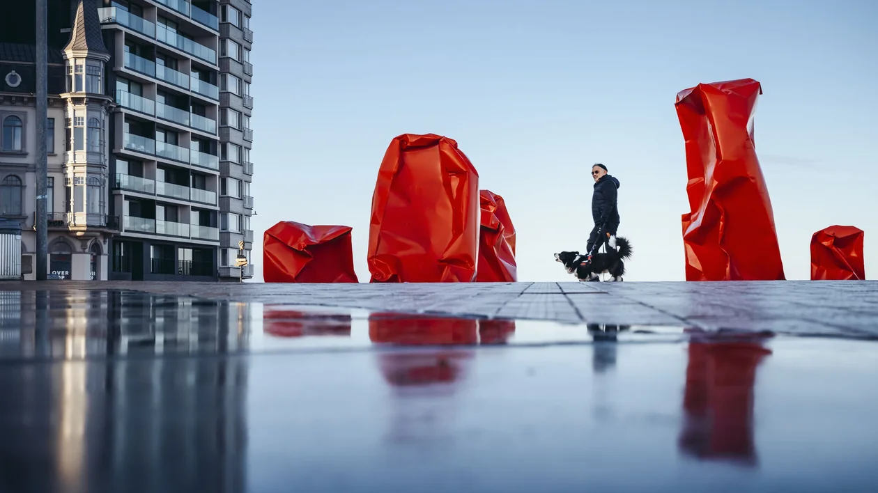 Person walking a dog past large red sculptures on a plaza with buildings in the background.