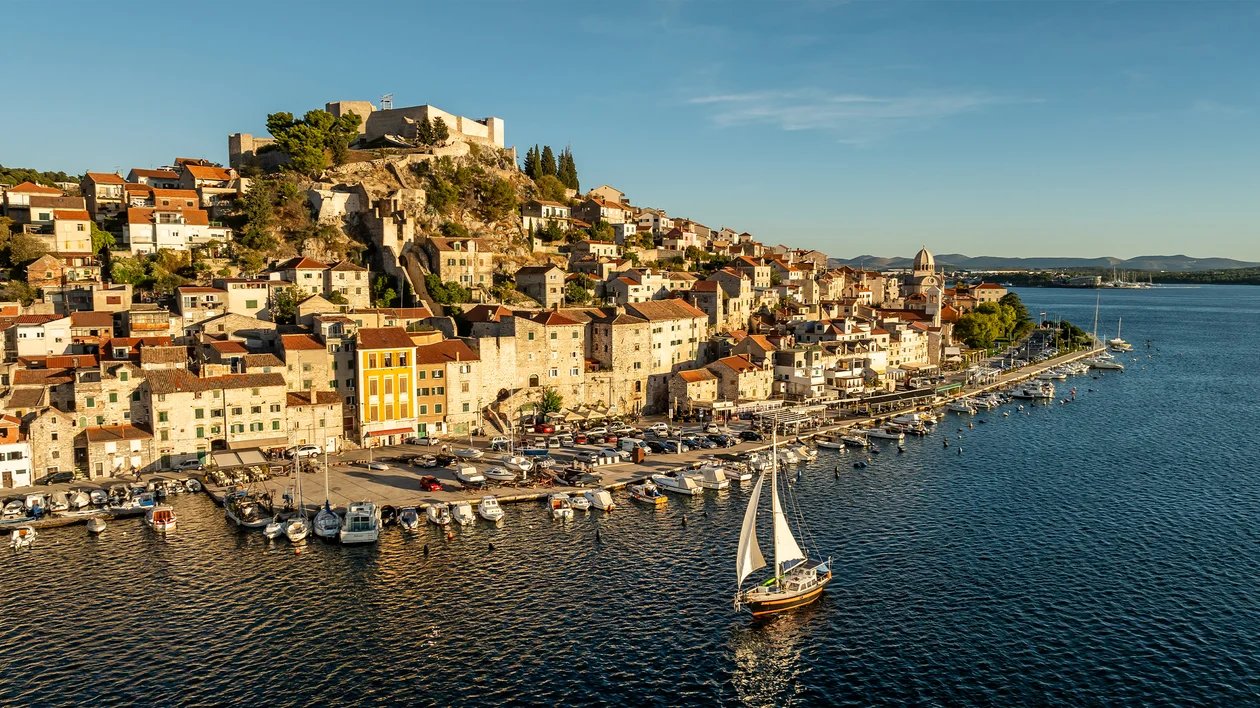 Sailboat near a coastal town with colorful houses and a hilltop fortress.