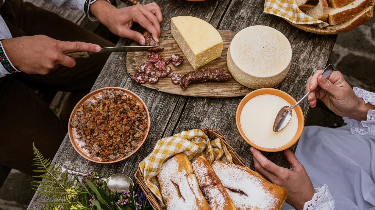 Rustic table with cheese, sausage, bread, pastries, granola, and two people eating.