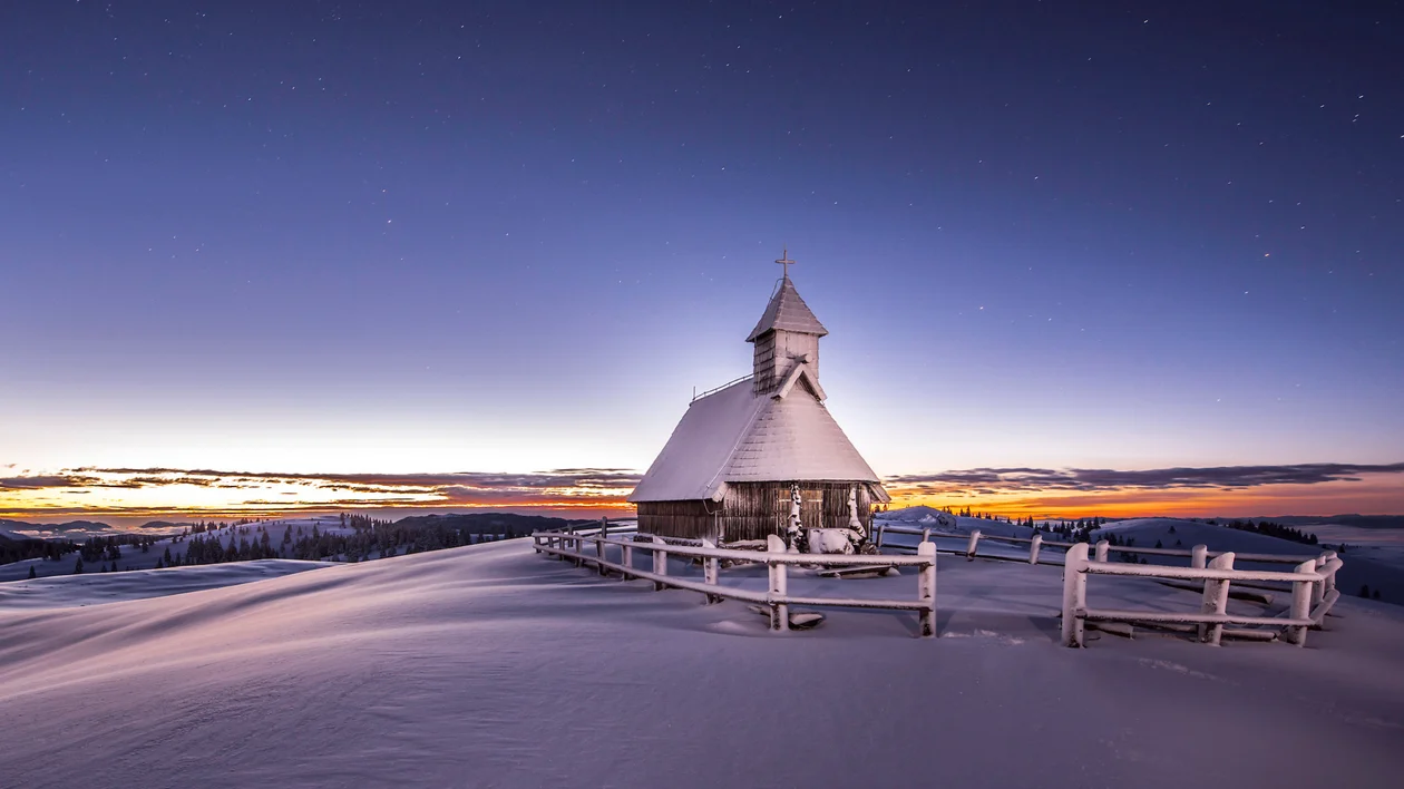Snow-covered church and fence at sunrise with a colorful sky.