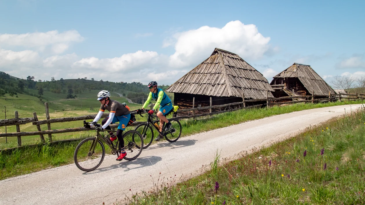 Two cyclists ride on a rural path past old wooden barns and green fields.